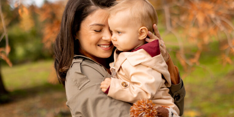 A woman smiles as she holds a baby close in a peaceful outdoor setting.